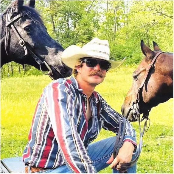 John Combs in a straw cowboy hat and sunglasses, sitting between two horses in a green field